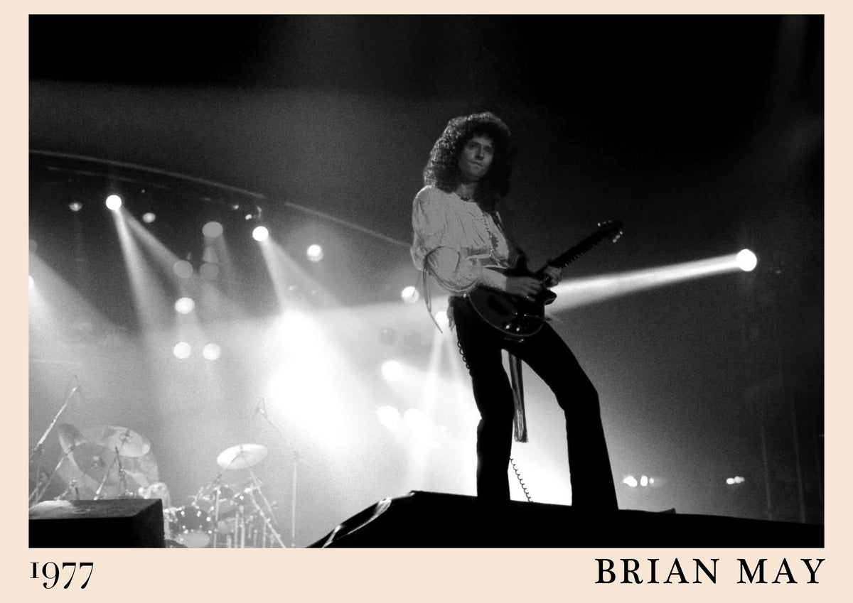 Black and white photo of Brian May playing guitar on stage under bright spotlights, with a drum kit behind him