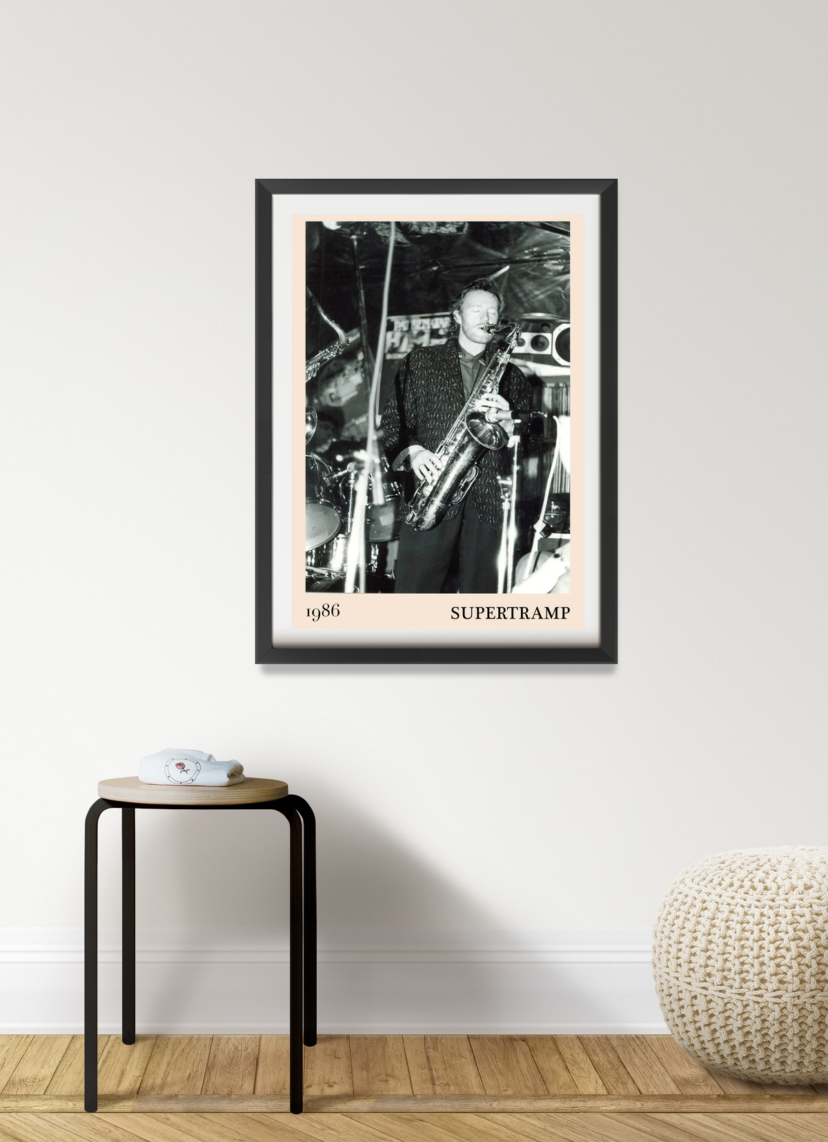 Framed Supertramp 1986 live concert print in black frame with pale pink mount, hung on a white wall above a small stool