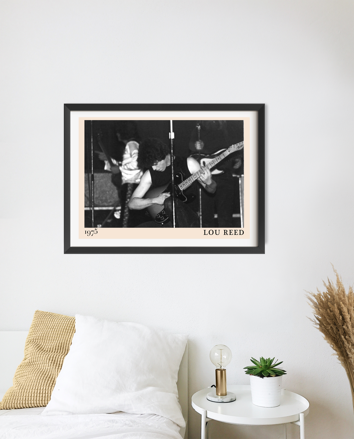 Framed Lou Reed 1975 concert print hung above a bedside table, featuring a black and white photo of guitarists performing on