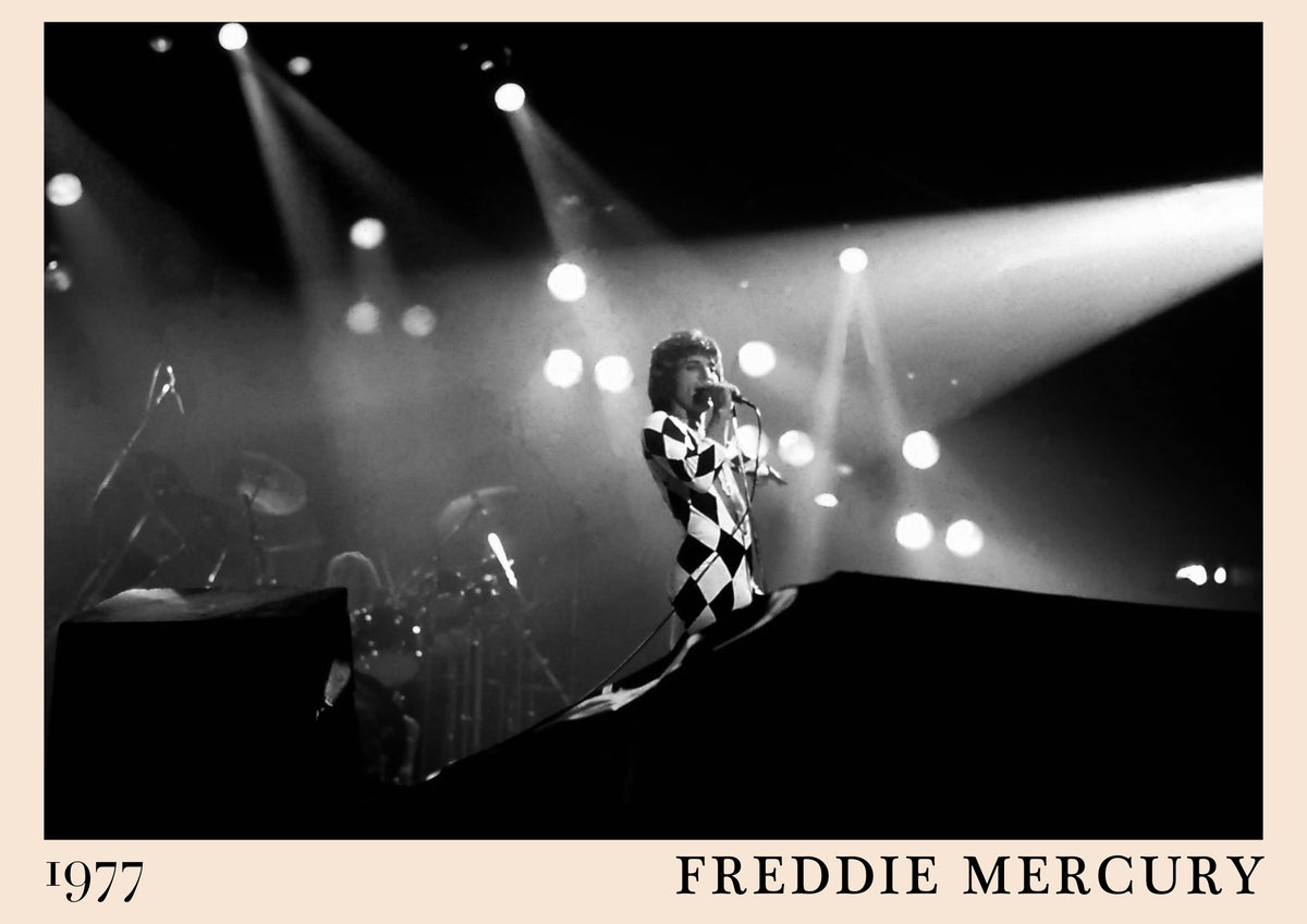 Black and white photo of Freddie Mercury singing on stage in a harlequin catsuit under spotlights, captioned 1977