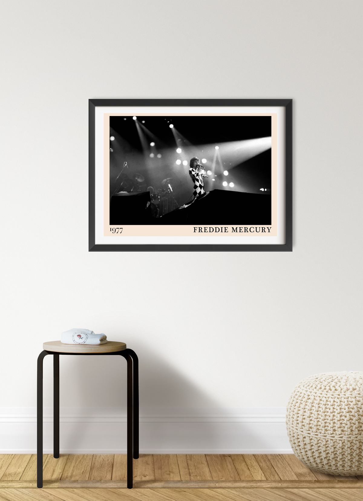 Freddie Mercury 1977 framed print in black frame with cream mount, hung on a white wall above a small stool and knitted