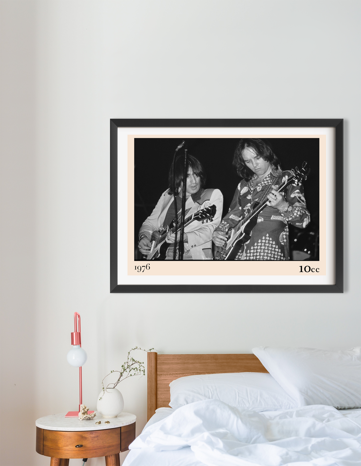 Framed 10cc 1976 black and white concert print hung above a wooden bed with white bedding and a round bedside table.