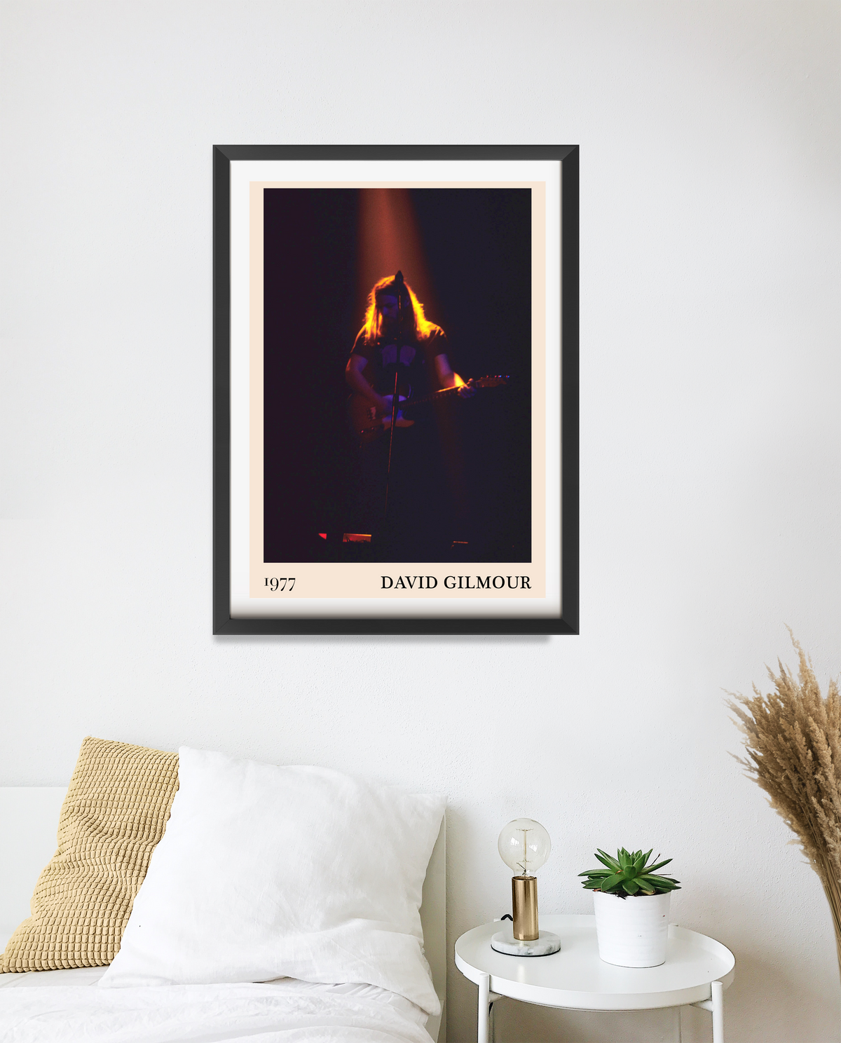 David Gilmour 1977 framed print in black frame, hung above a white bed with a bedside table holding a lamp and succulent.