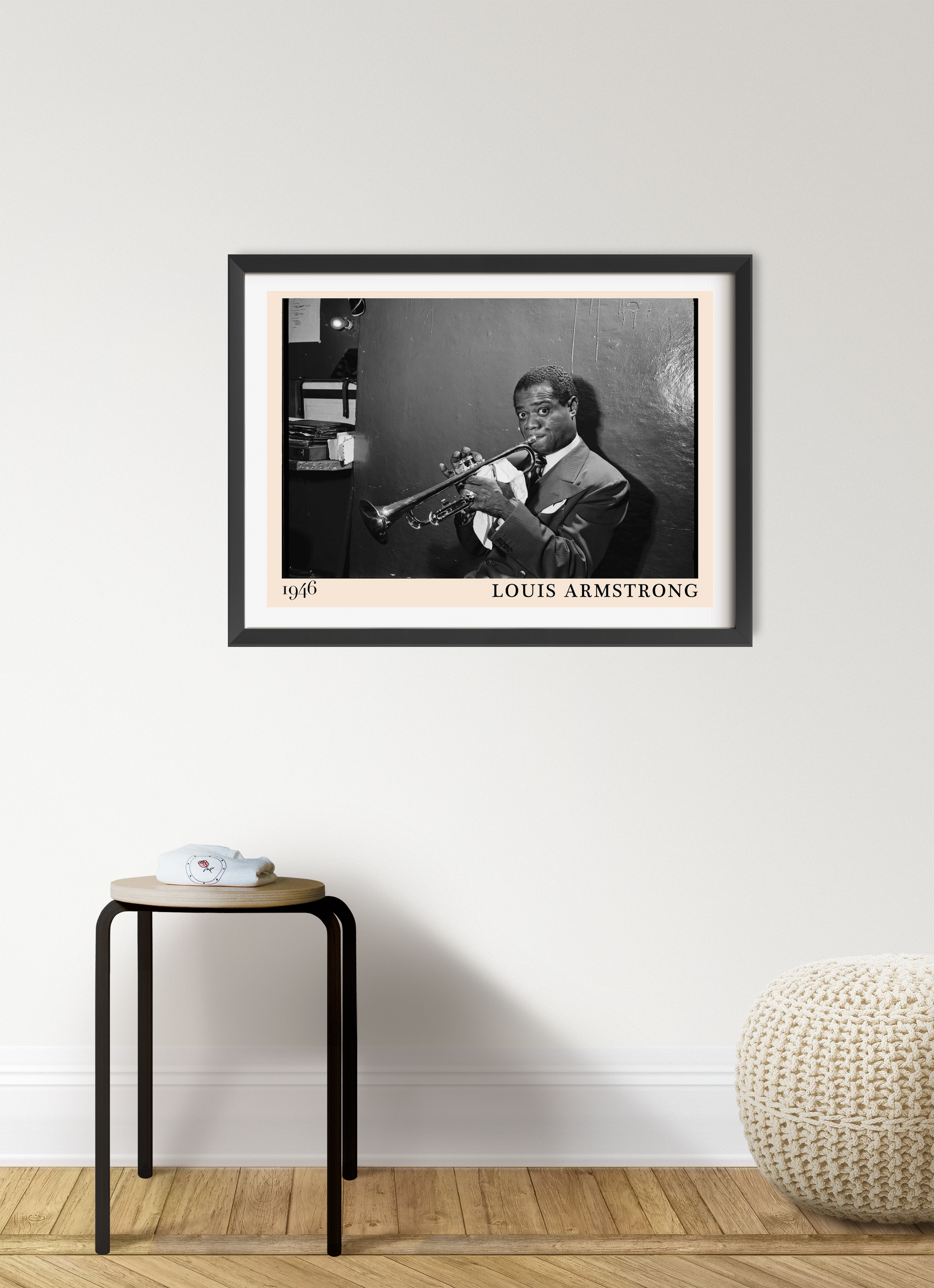 Louis Armstrong vintage jazz poster in a black frame on a modern white wall above a minimalist stool and knitted pouf.