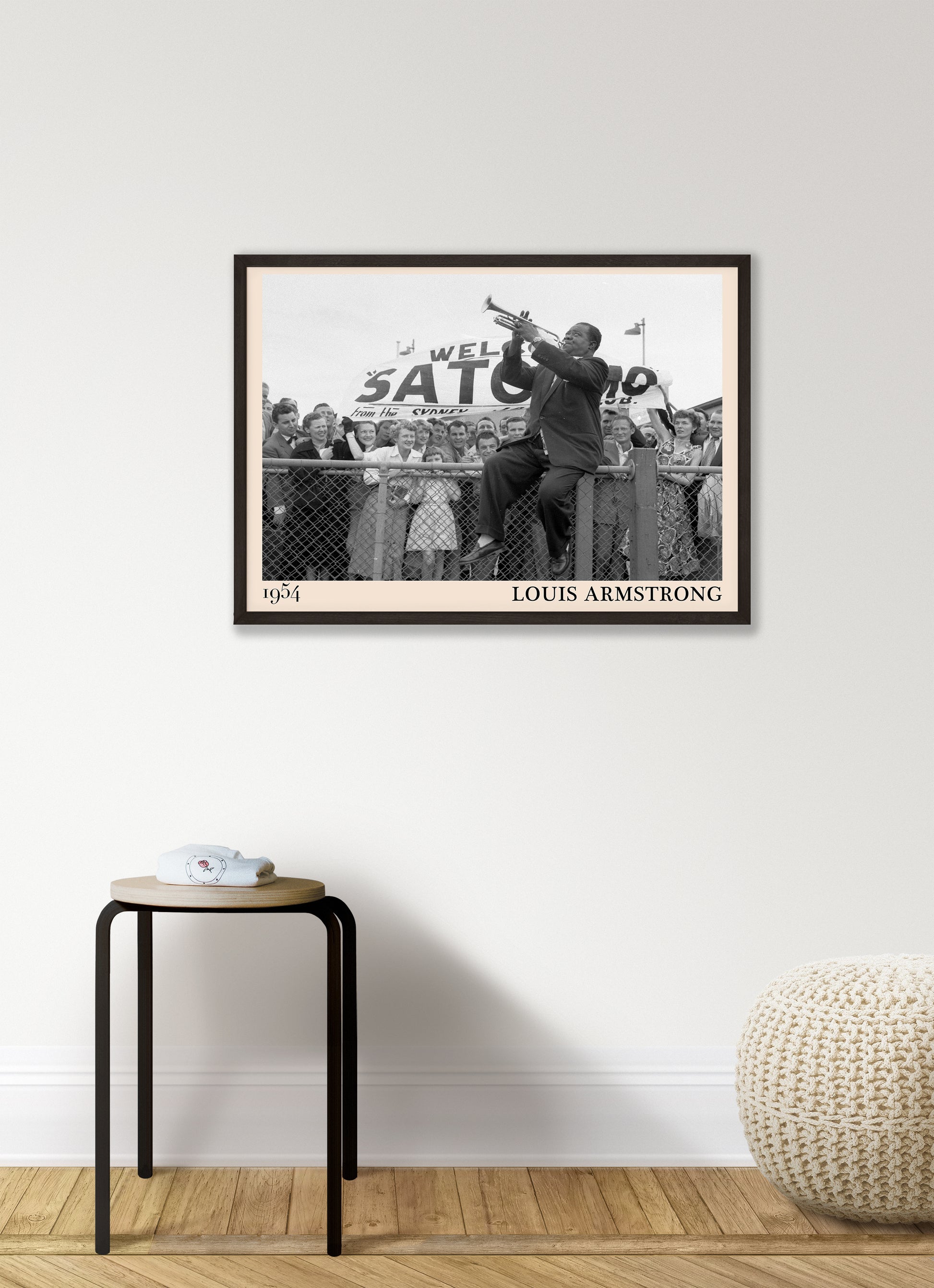 1954 picture of Louis Armstrong playing his trumpet sat on a fence. Picture crafted into a retro black framed jazz print, with an off-white border. Poster is hanging on a grey living wall