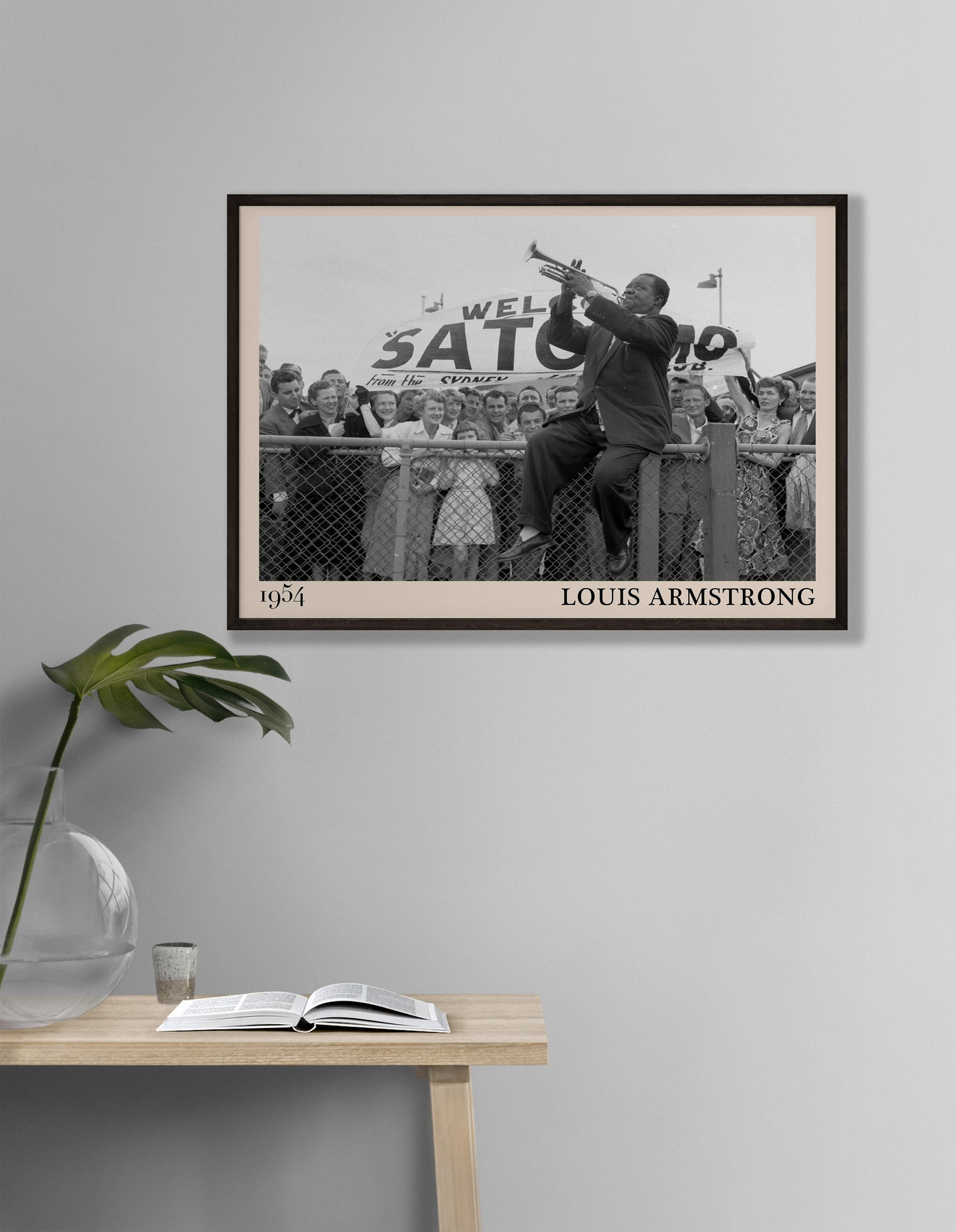 1954 picture of Louis Armstrong playing his trumpet sat on a fence. Picture crafted into a cool black framed jazz print, with an off-white border. Poster is hanging on a grey living room wall