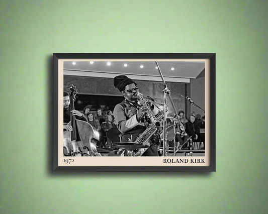 1972 photo of Roland Kirk crafted into a black framed-poster, hanging on a green coloured concrete wall in dappled sunlight