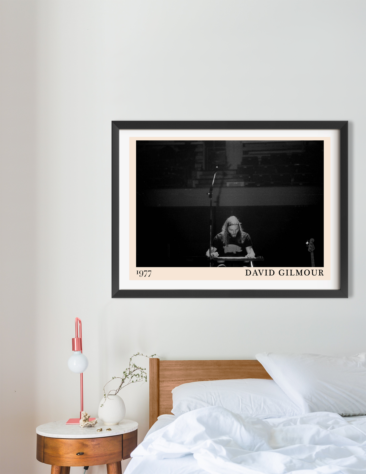 Framed David Gilmour 1977 concert print in black frame with cream mount, hung above a wooden bed in a minimal bedroom.