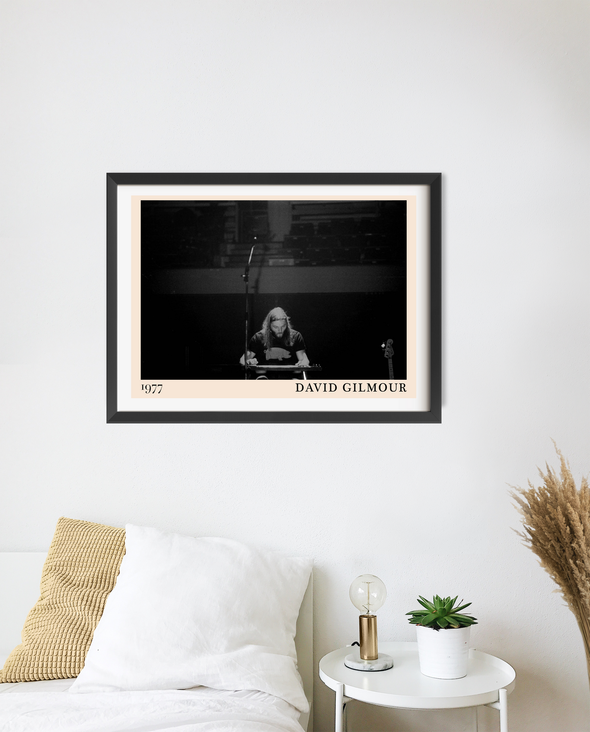 Framed David Gilmour 1977 concert print in black frame, hung above a bedside table with a lamp and small plant.