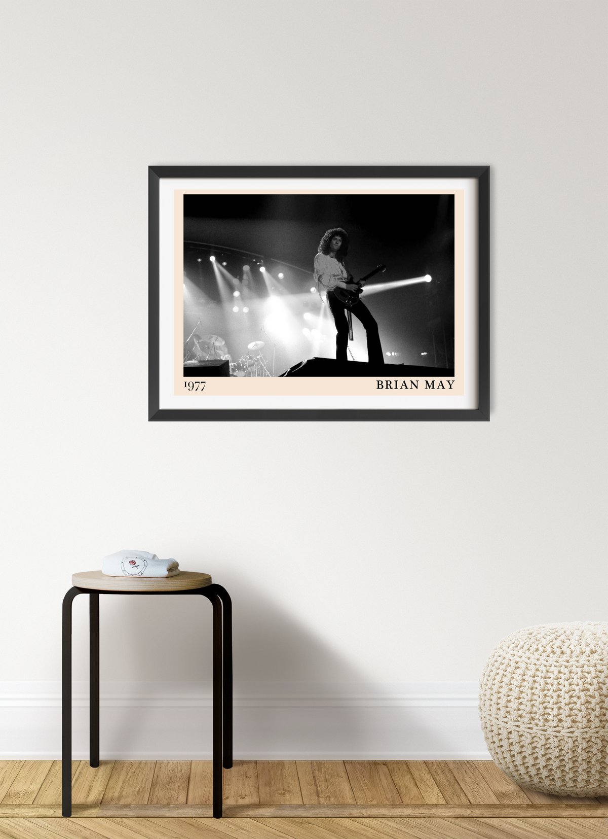 Framed print of Brian May performing on stage in 1977, shown in black and white, hung above a wooden stool on a white wall.