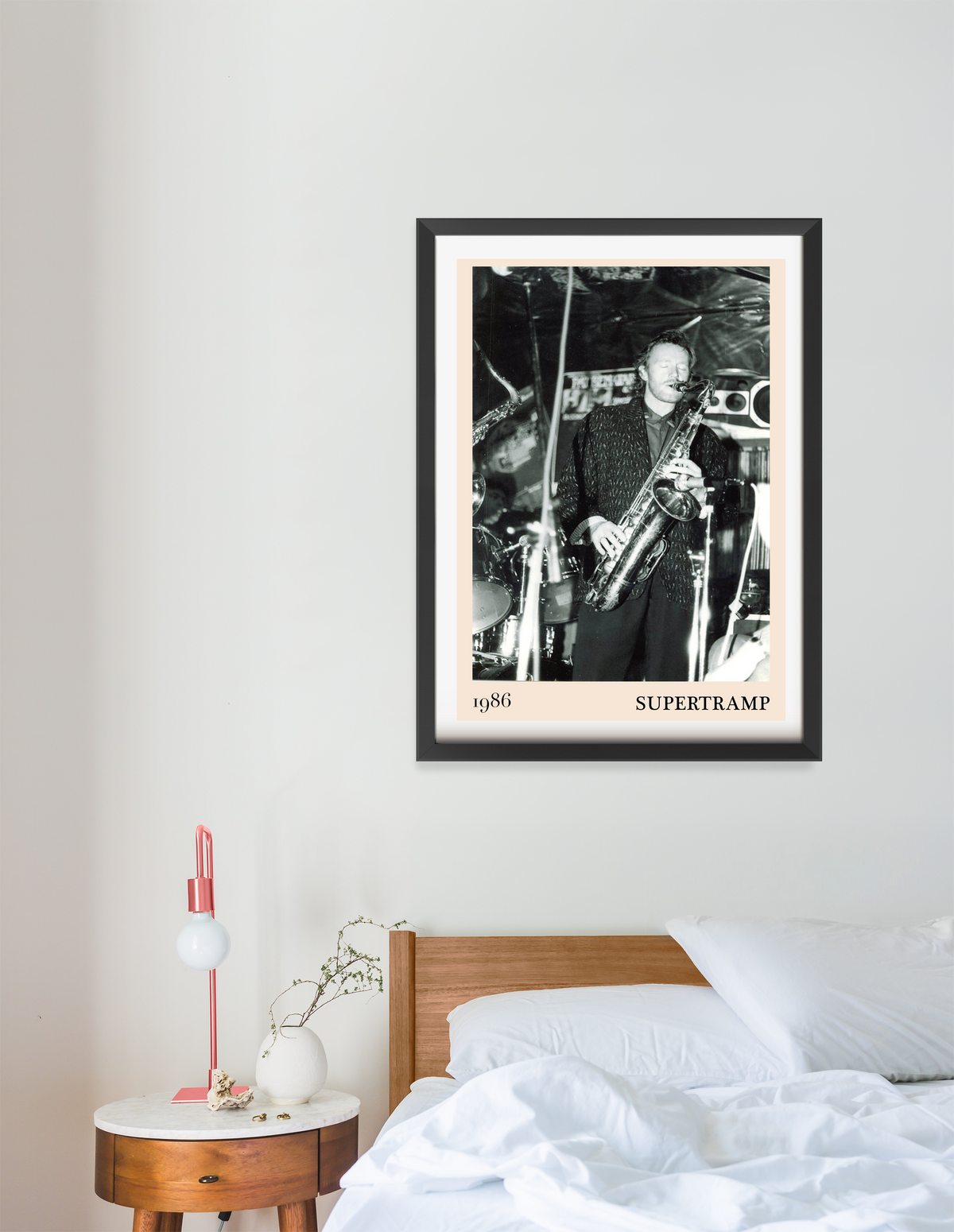 Framed Supertramp 1986 concert print in black frame hanging above a wooden bed with white bedding and a bedside table.