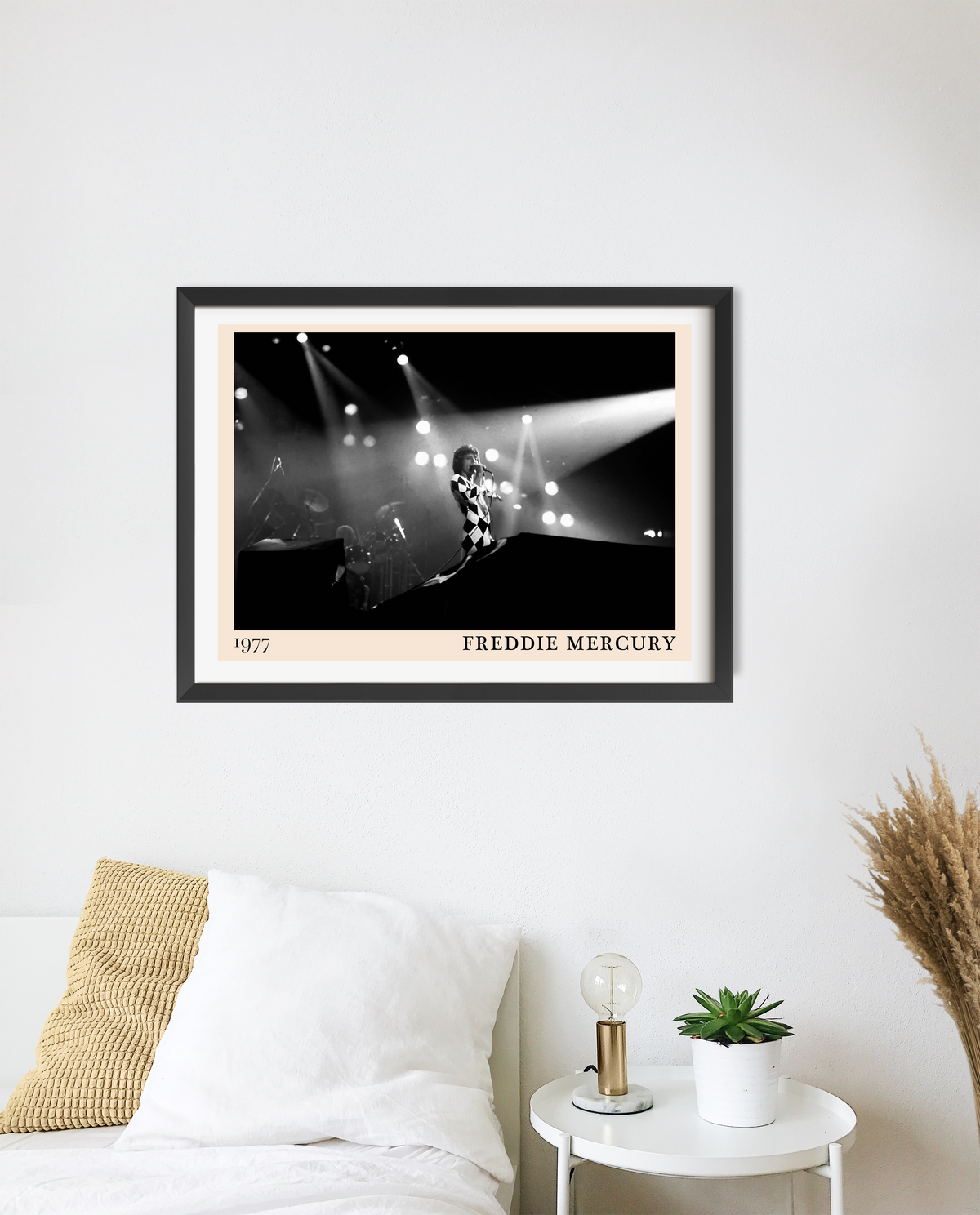 Freddie Mercury 1977 framed print in black frame with cream mount, hung above a bedside table with a lamp and succulent.